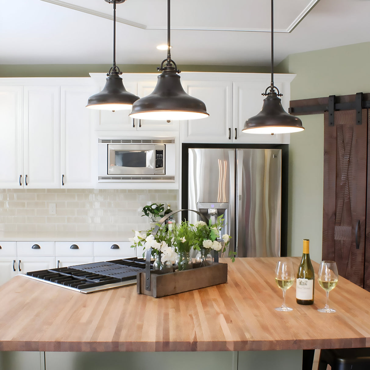 A modern kitchen showcases white cabinets, stainless steel appliances, and a wooden island. Overhead, three vintage-inspired Emery 1 Light Medium Pendants in Palladian Bronze emit a warm glow on the decorative flower arrangement. Two glasses and a wine bottle are invitingly displayed on the counter.