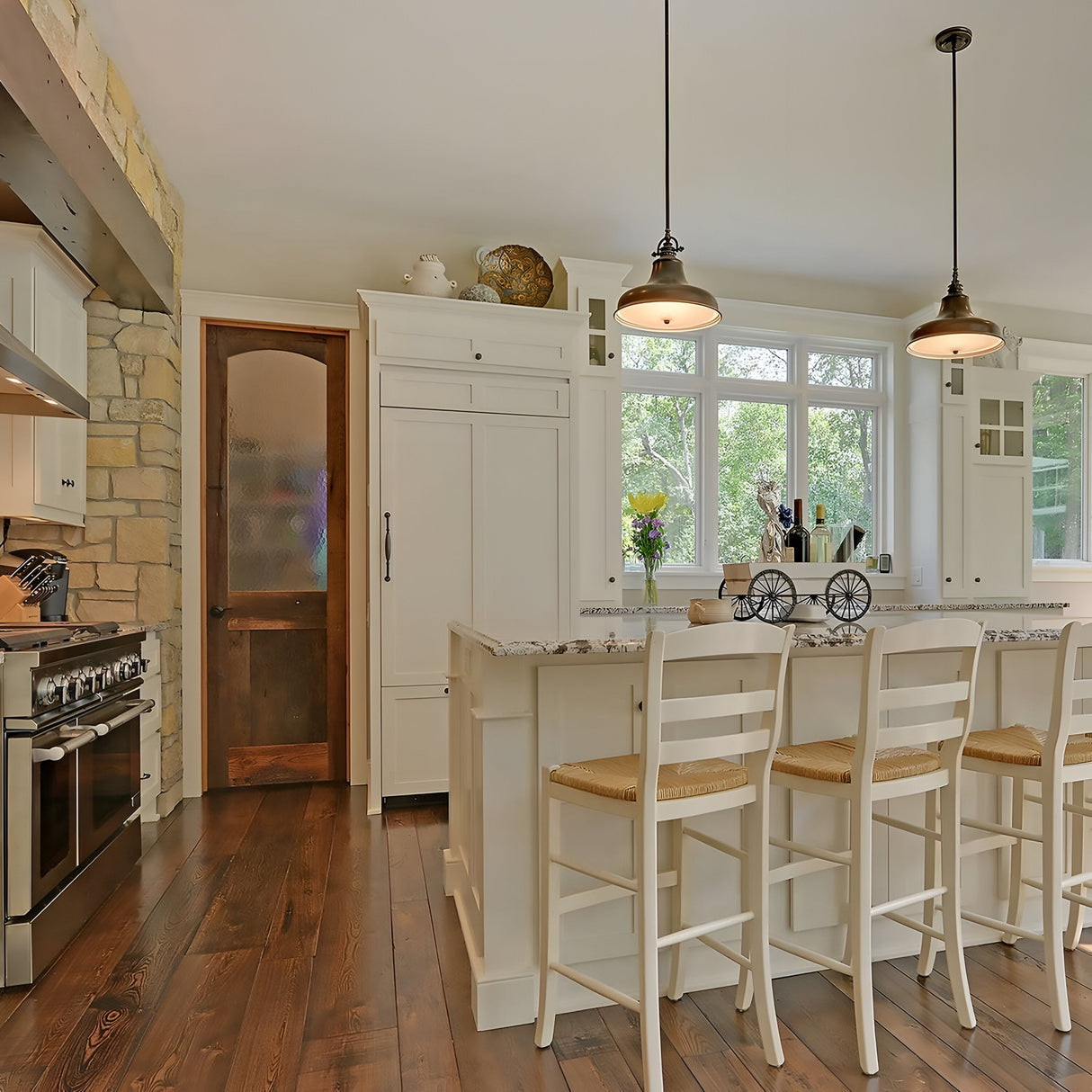 The kitchen is cozy, featuring hardwood floors, white cabinets, and a spacious island surrounded by four wooden chairs. Above the island, Emery 1 Light Medium Pendant fixtures in weathered brass provide warm lighting while large windows allow natural light to flood in. To the left is a door with a frosted glass panel.