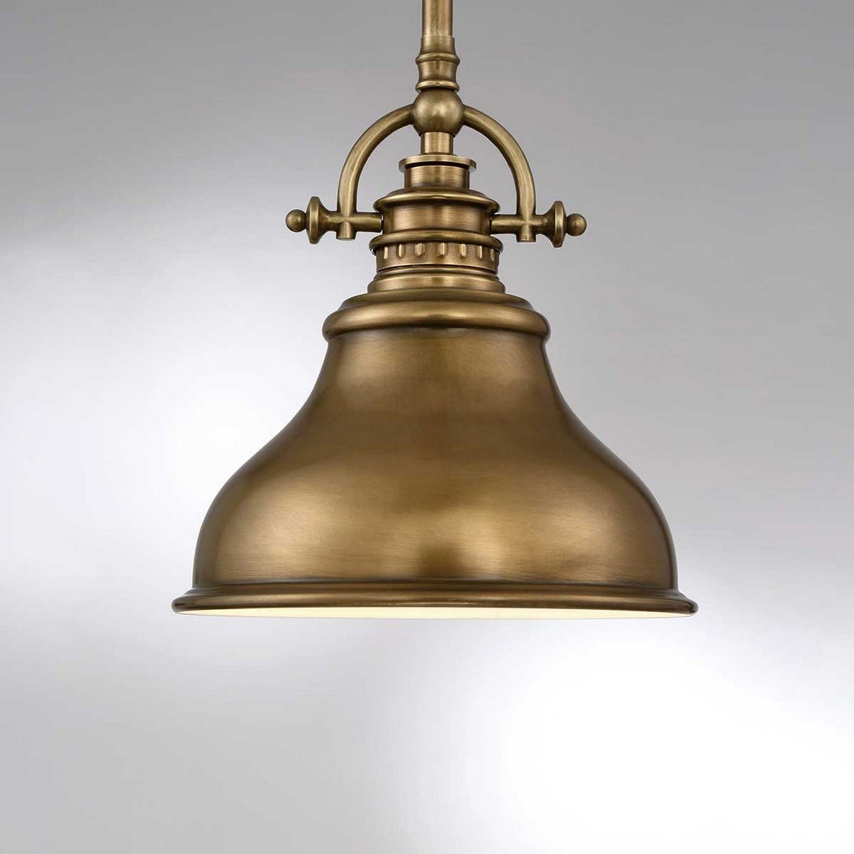 The Emery 1 Light Mini Pendant in Weathered Brass, featuring a bell-shaped shade, is suspended elegantly from a round ceiling mount. Its metallic surface has a smooth matte finish that emits a warm glow, perfectly complemented by the plain gray backdrop that enhances its timeless charm.