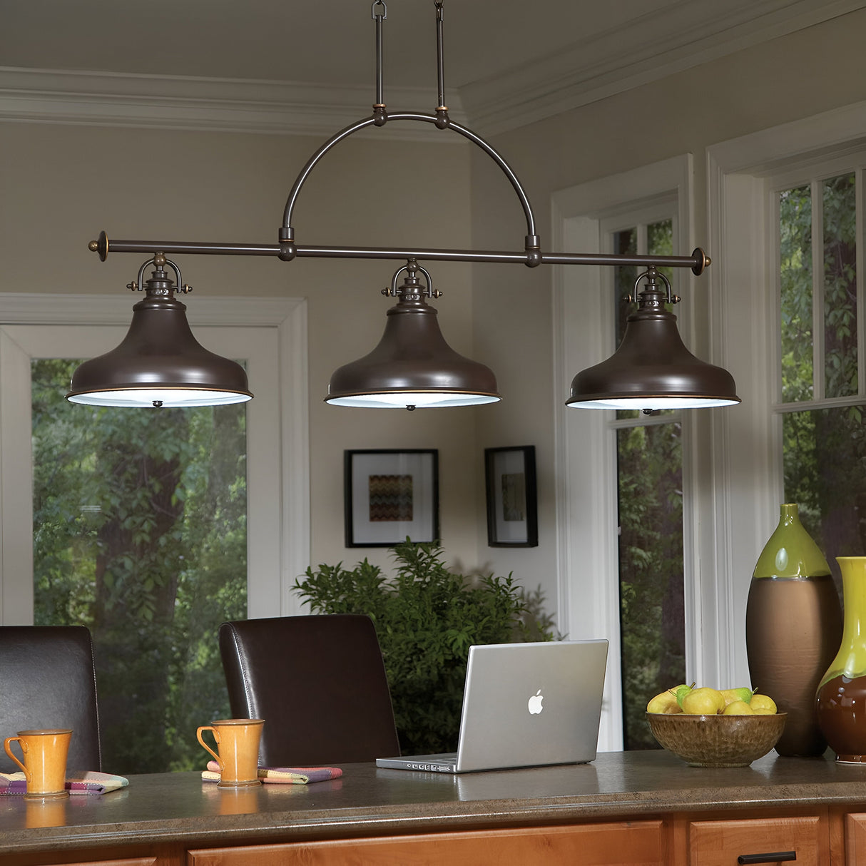A kitchen with rustic charm features Palladian Bronze Emery 3 Light Island Pendants illuminating a wooden counter. A laptop, two coffee mugs, a green vase, and a bowl of fruit are neatly arranged. Large windows and framed art provide a serene backdrop.