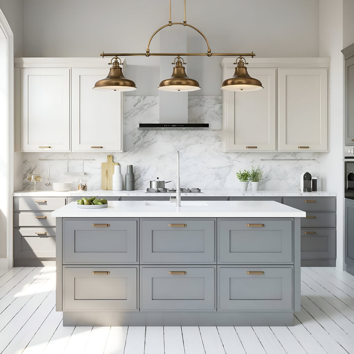The modern kitchen is equipped with white and gray cabinets, a marble backsplash, and a spacious central island. Above the island are three Emery 3 Light Island Pendants in Weathered Brass, which add a charming touch. The room showcases a minimalist design, enhanced by natural light streaming through large windows.