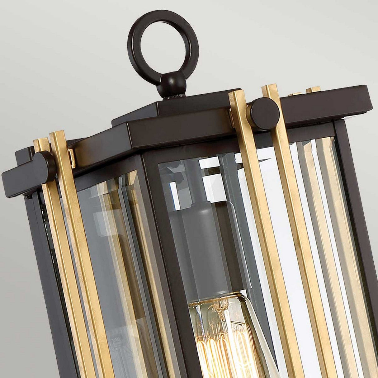 A close-up of the Goldenrod Outdoor 1 Light Medium Wall Lantern showcases its contemporary design with a bronze metal frame and vertical gold detailing. Clear glass panels display an Edison-style bulb within, while a circular top handle enhances its decorative appeal. Its style subtly nods to mid-century lighting, set against a neutral off-white background.