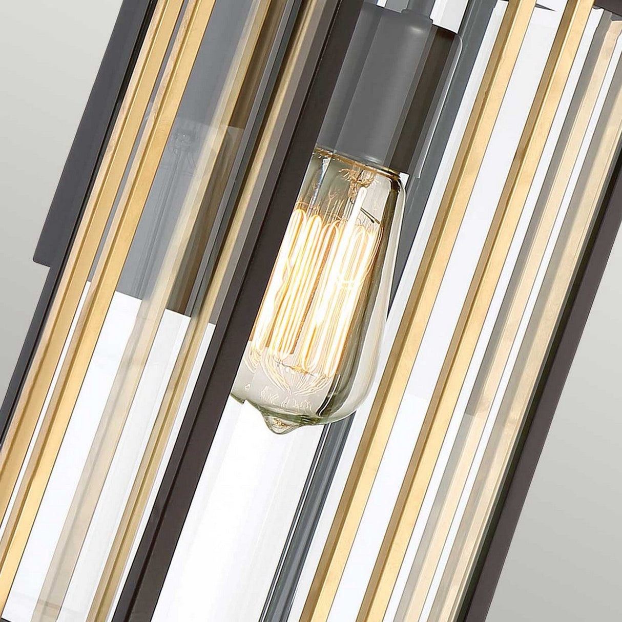 Close-up of the Goldenrod Outdoor 1 Light Medium Wall Lantern - Bronze, showcasing an exposed Edison bulb encased by sleek metal bars that evoke a Mid-Century Lighting style. The contrasting gold and black bars harmonize with clear glass panels, offering a sophisticated industrial aesthetic.