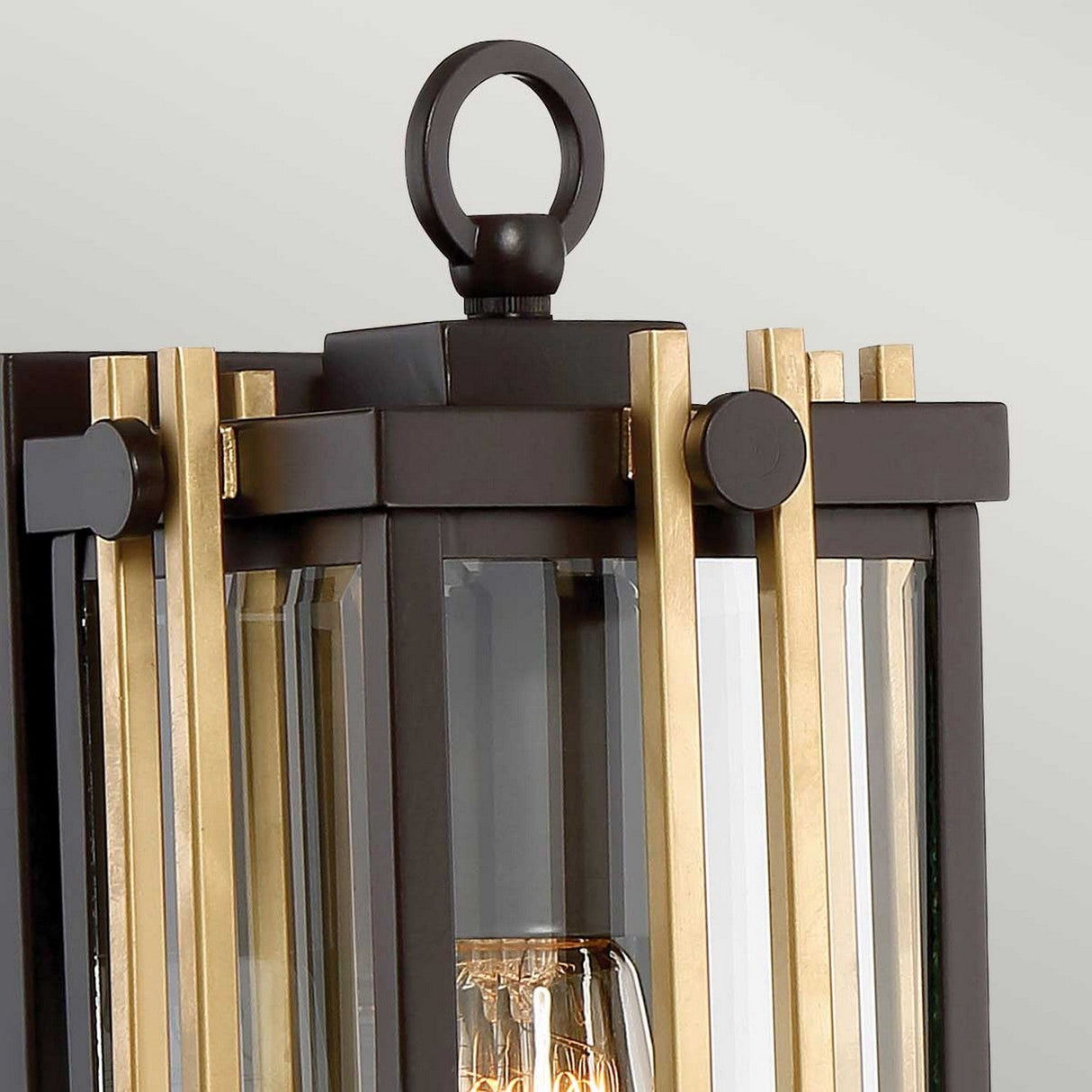 Close-up of the Goldenrod Outdoor 1 Light Small Wall Lantern in a Western bronze finish, showcasing its modern geometric design. It features black and gold metal bars surrounding bevelled glass panels, with a visible bulb inside, and has a circular loop on top for hanging.