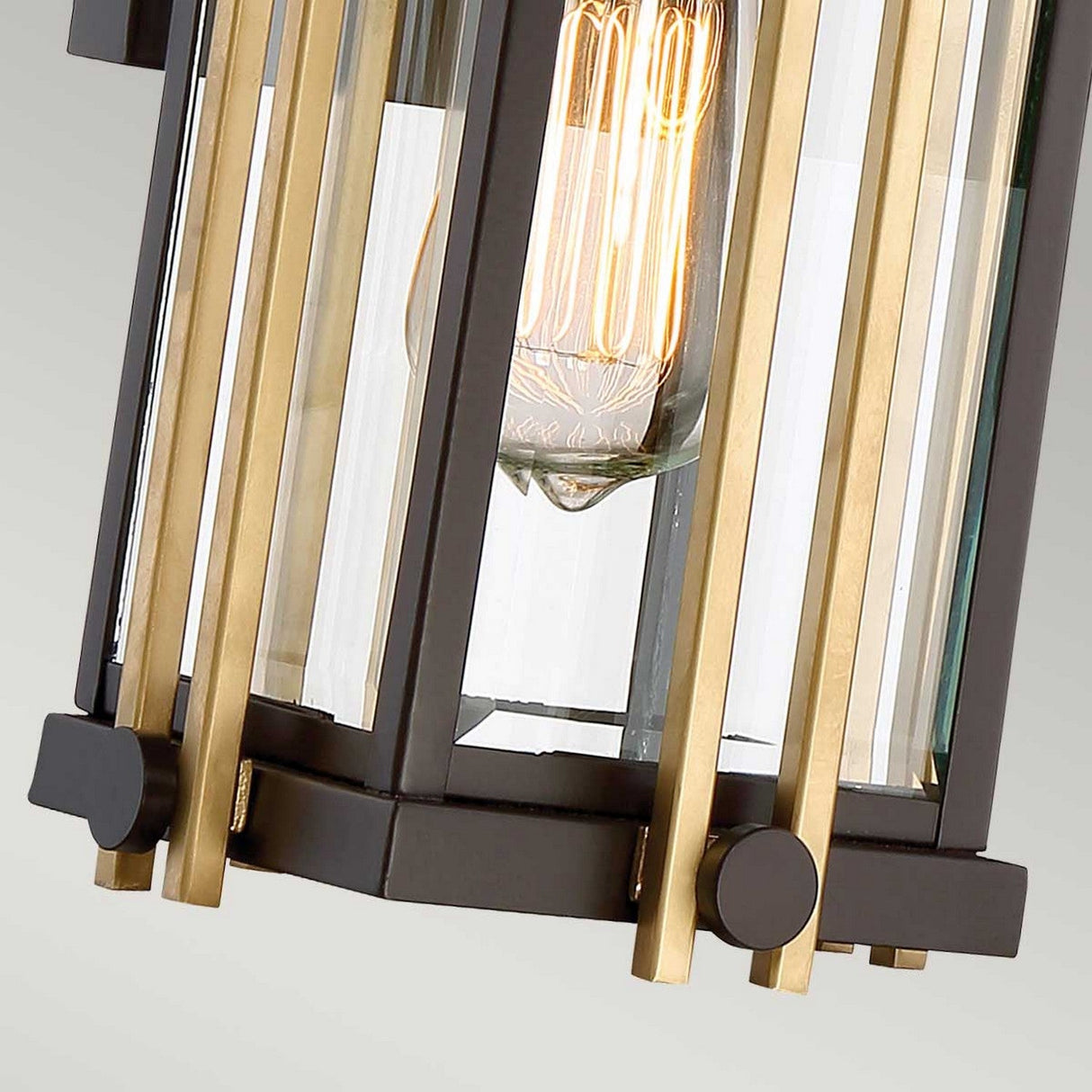 A close-up of the Goldenrod Outdoor 1 Light Small Wall Lantern in bronze showcases its contemporary design, featuring vertical gold and black metal bars complemented by beveled glass panels. The exposed Edison bulb inside emits a warm glow, enhancing its modern and elegant aesthetic against a plain, soft grey background.
