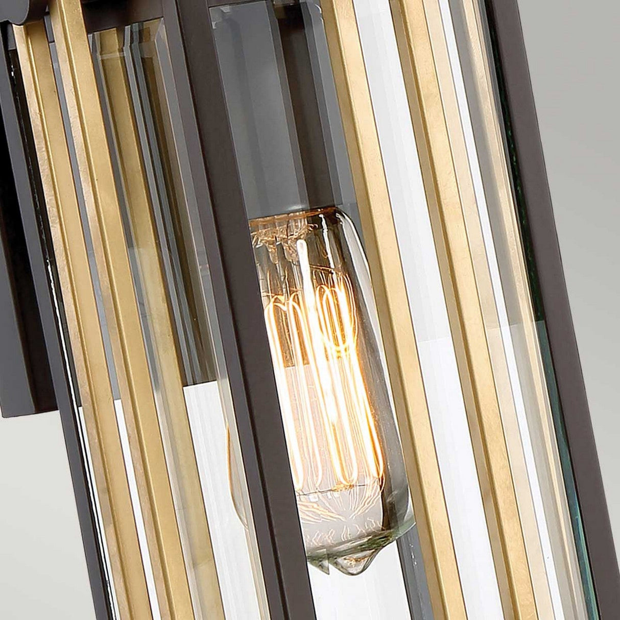 A close-up of the Goldenrod Outdoor 1 Light Small Wall Lantern - Bronze showcases a modern pendant light fixture with an exposed filament bulb encased in clear and bevelled glass panels, emitting a warm glow. The subtle hints of gold accentuate the design similarly to the elegant Western Bronze Finish.