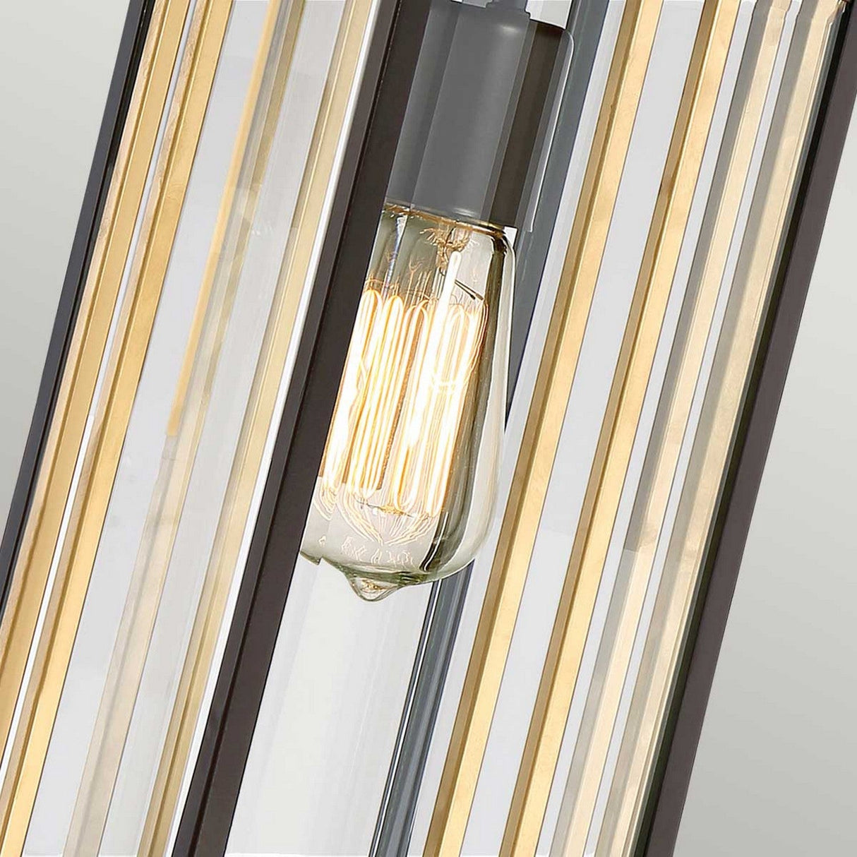 Close-up of the Goldenrod Outdoor 1 Light Medium Chain Pendant in a Western Bronze finish, featuring an exposed Edison bulb. The bulb emits a warm glow, encased by vertical glass panels that contribute to an elegant mid-century style against a neutral background.