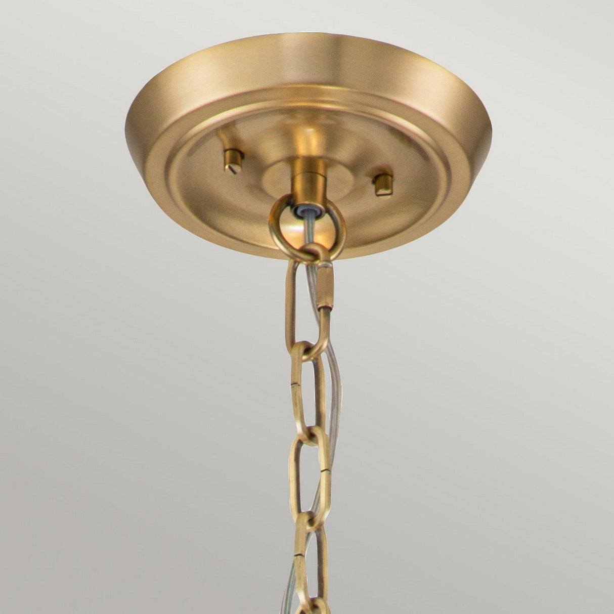 Close-up of a Gotham 4 Light Small Chandelier in a brushed brass finish, showcasing its hanging chain and circular ceiling mount. The fixture, adorned with an elegant cream shade, is displayed against a light gray background.
