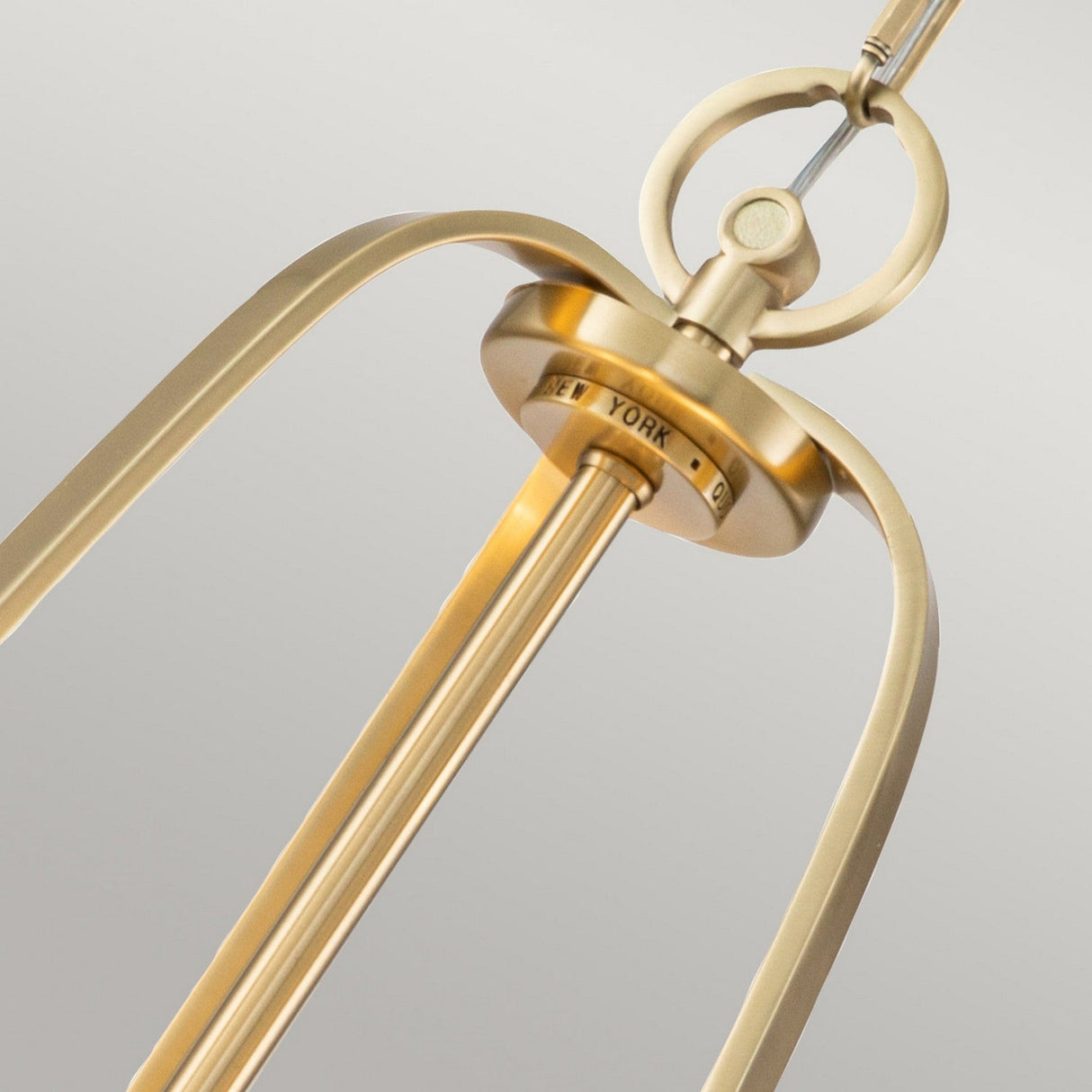 Close-up of a Gotham 4 Light Small Chandelier in brushed brass with a contemporary golden pendant light fixture, showcasing three elegant rods and a circular mounting element. The central piece is engraved with the words NEW YORK, all set against a neutral background.