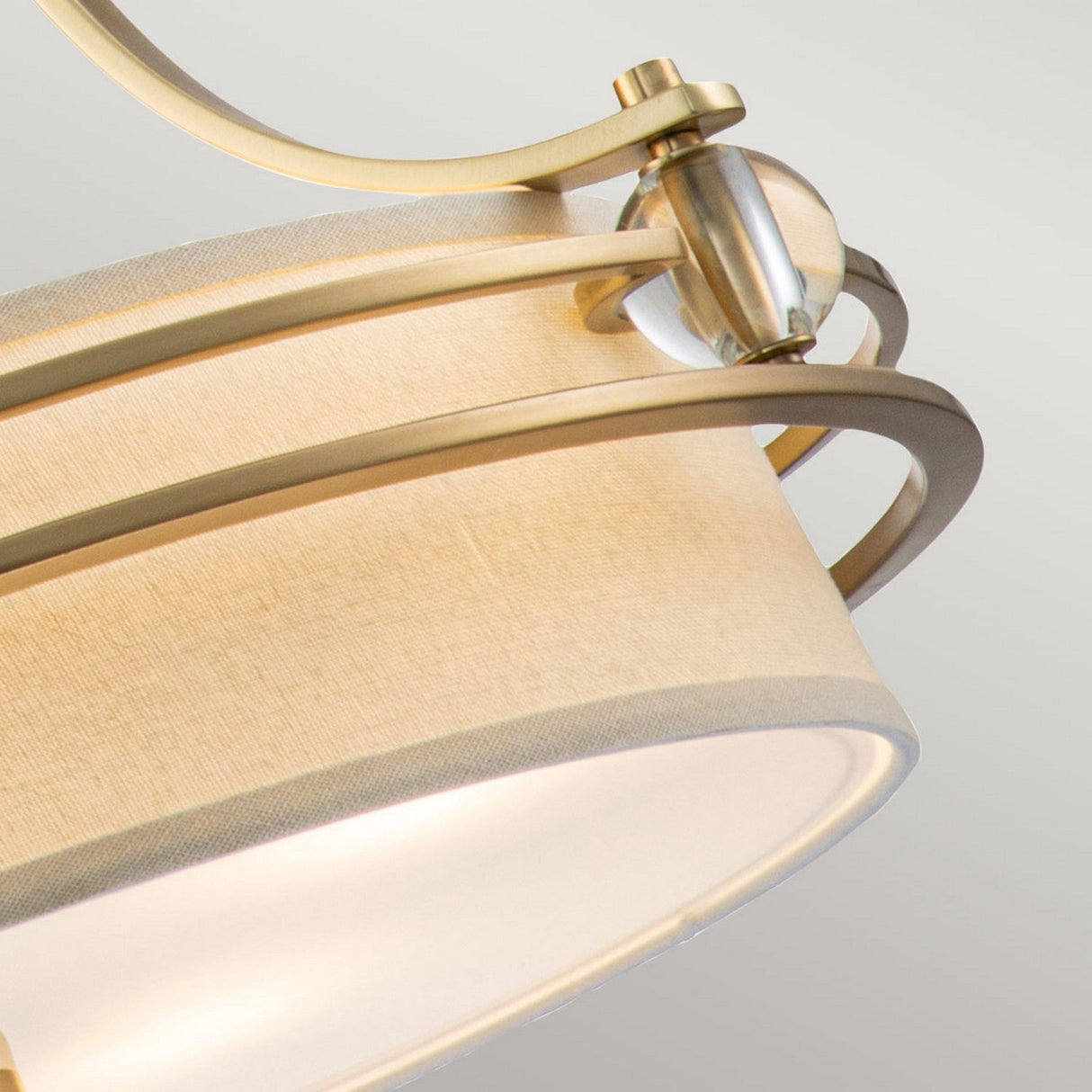 A close-up view of a Gotham 4 Light Small Chandelier in brushed brass showcases a modern lamp with a cream fabric shade and elegant accents. The design includes curved metal arms that hold the shade, echoing the chandelier's sophisticated allure against a neutral gray background.