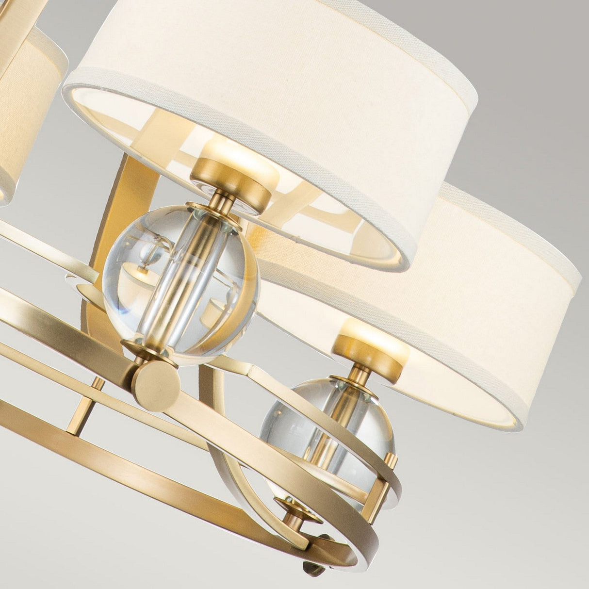 A close-up of the Gotham 4 Light Chandelier in brushed brass showcases urban-inspired elegance with its four drum-shaped, cream-colored fabric lampshades. The fixture's brushed brass frame harmoniously complements its spherical glass accents, set against a neutral gray background.