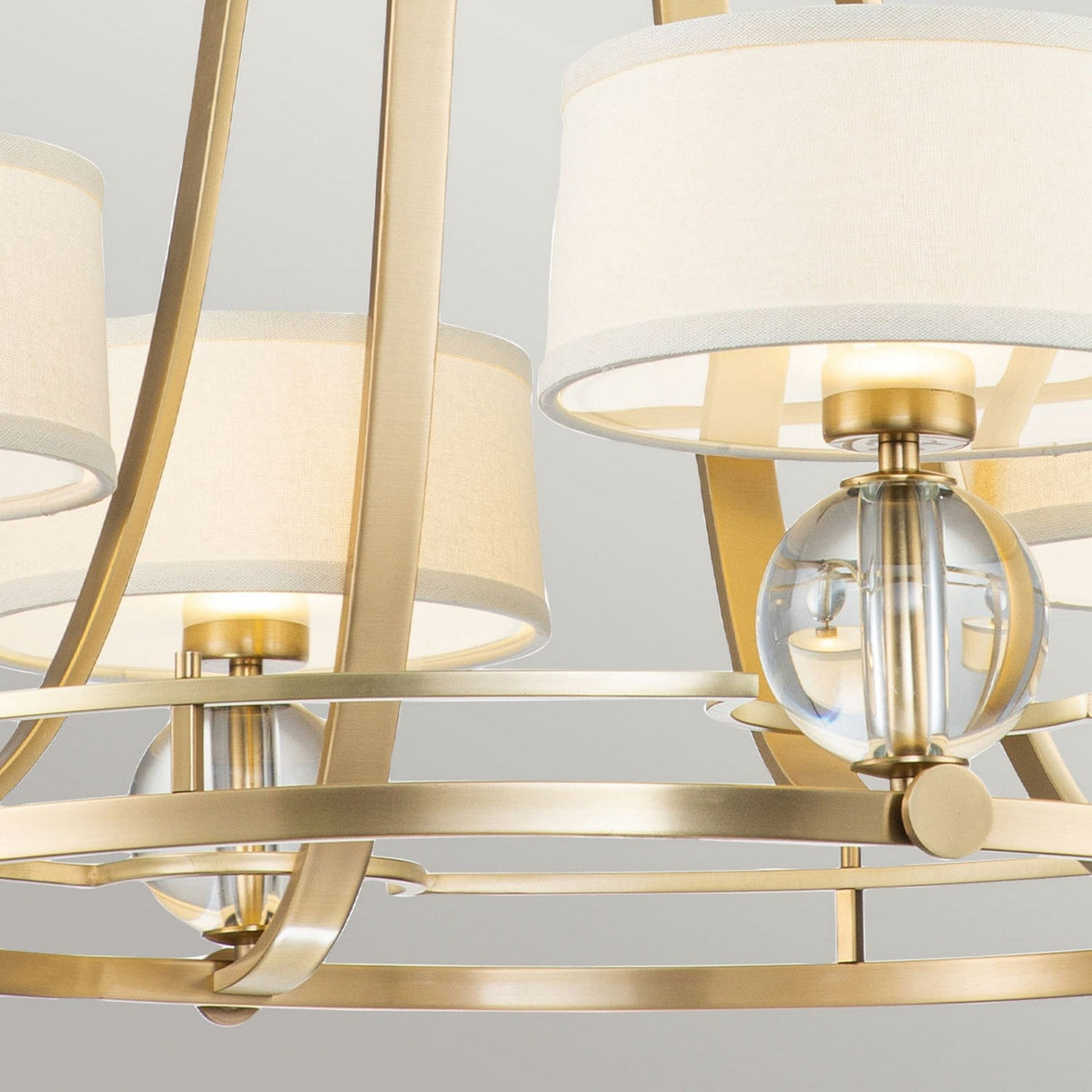 Close-up of the Gotham 4 Light Chandelier - Brushed Brass, showcasing a gold metal frame and multiple white drum lampshades. The design, with circular glass accents and graceful curved lines, embodies urban-inspired elegance for a sophisticated, contemporary look.