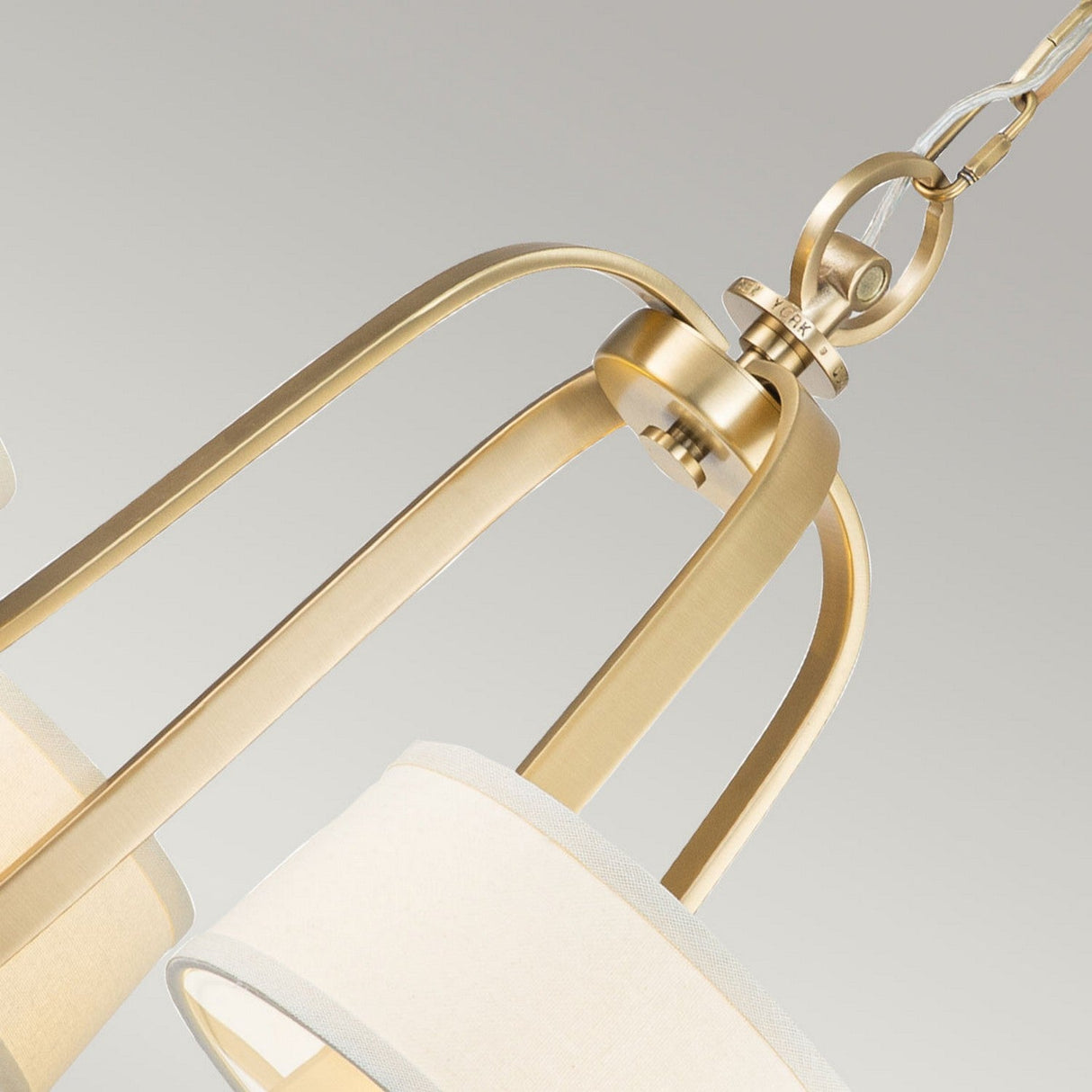 The Gotham 4 Light Chandelier - Brushed Brass exudes urban-inspired elegance with its modern gold pendant light fixture. It features brushed brass curved metal bars and a chain link that beautifully complement the cream-colored cylindrical fabric shades, adding a soft touch to the sophisticated design.