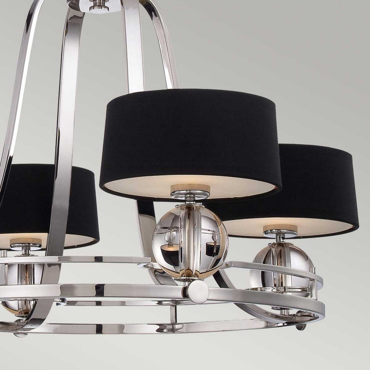 The Gotham 4 Light Chandelier - Imperial Silver exudes industrial elegance with its metallic highlights and sleek chrome finish against a neutral gray background, and it features four evenly spaced lights complemented by black drum lampshades for a touch of sophistication.