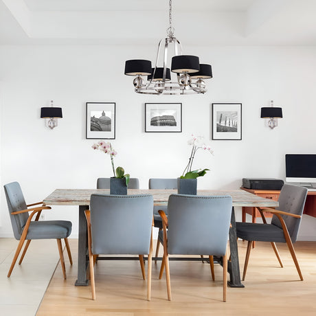 A modern dining room showcases a wooden table surrounded by six gray chairs. The Gotham 4 Light Chandelier in Imperial Silver elegantly hangs above, casting a sophisticated light. Orchids adorn the table, while black-and-white photos enhance the walls' industrial elegance, complementing the corner desk with a computer.