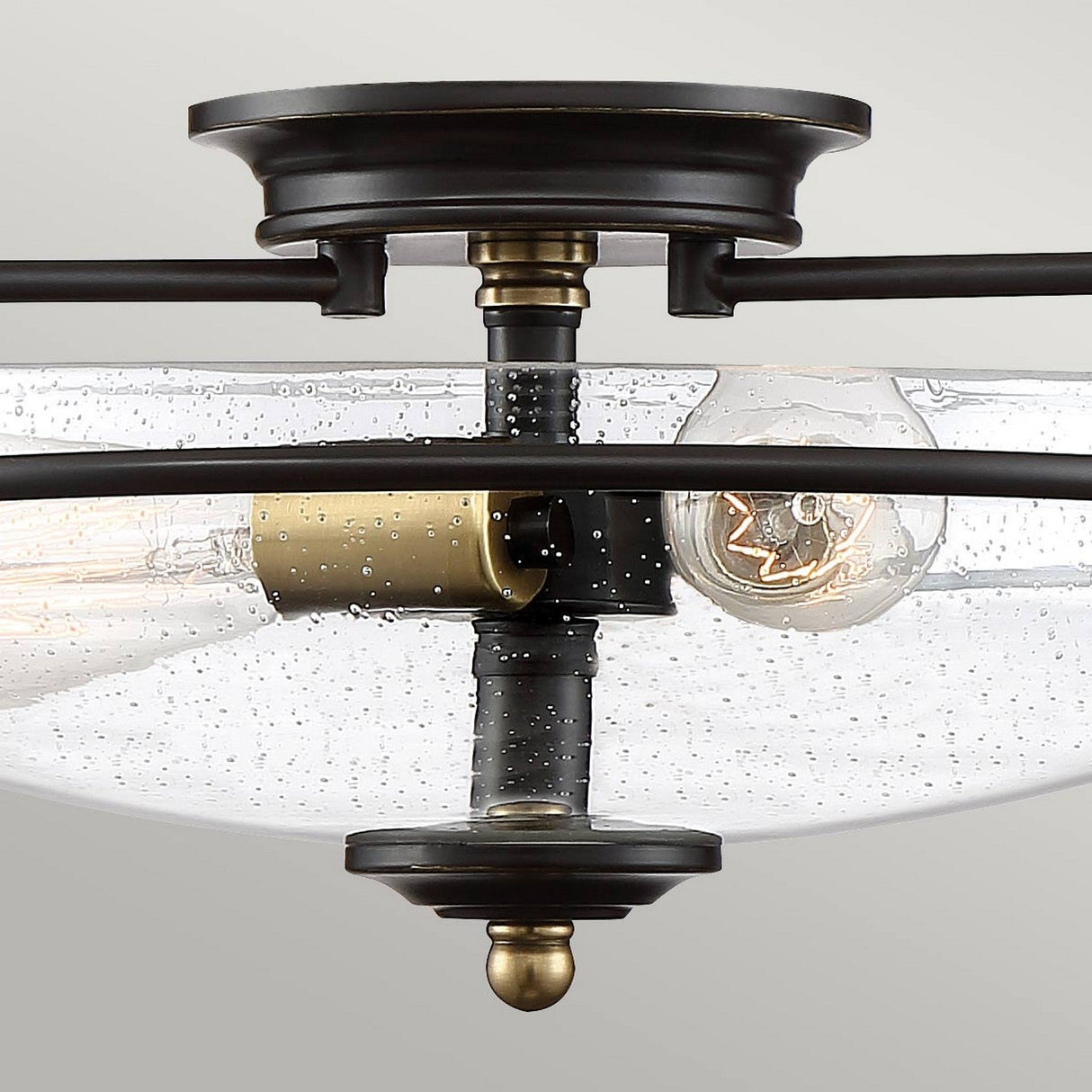 Close-up of a Palladian Bronze and weathered brass semi-flush mount ceiling light fixture from the Griffin collection, featuring a clear, seeded glass shade. This elegant fixture comes with three exposed light bulbs, adding a vintage touch to its modern design enhanced by the bubbled texture of the glass.