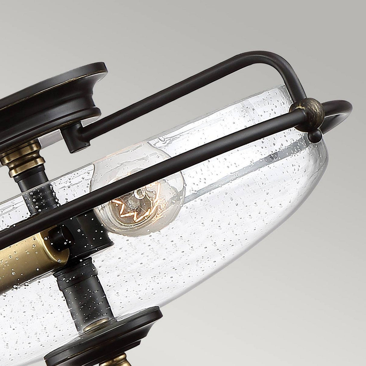 Close-up of the Griffin 3 Light Flush, showcasing a modern design with a clear, dome-shaped glass shade adorned with small air bubbles. Inside, an exposed Edison bulb is visible, encased in a dark Palladian Bronze frame with Weathered Brass accents. The background is light gray.