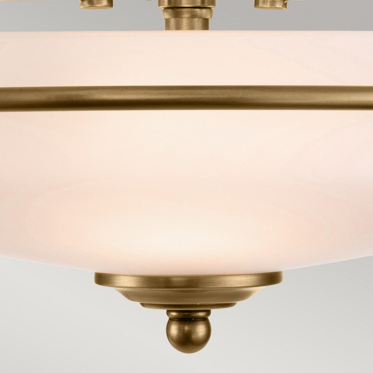 A close-up of the Griffin 3 Light Flush - Weathered Brass ceiling fixture showcases its round, frosted glass shade enhanced by weathered brass details and a decorative finial. Ideal for transitional interiors, it harmonizes beautifully with the neutral gray backdrop.