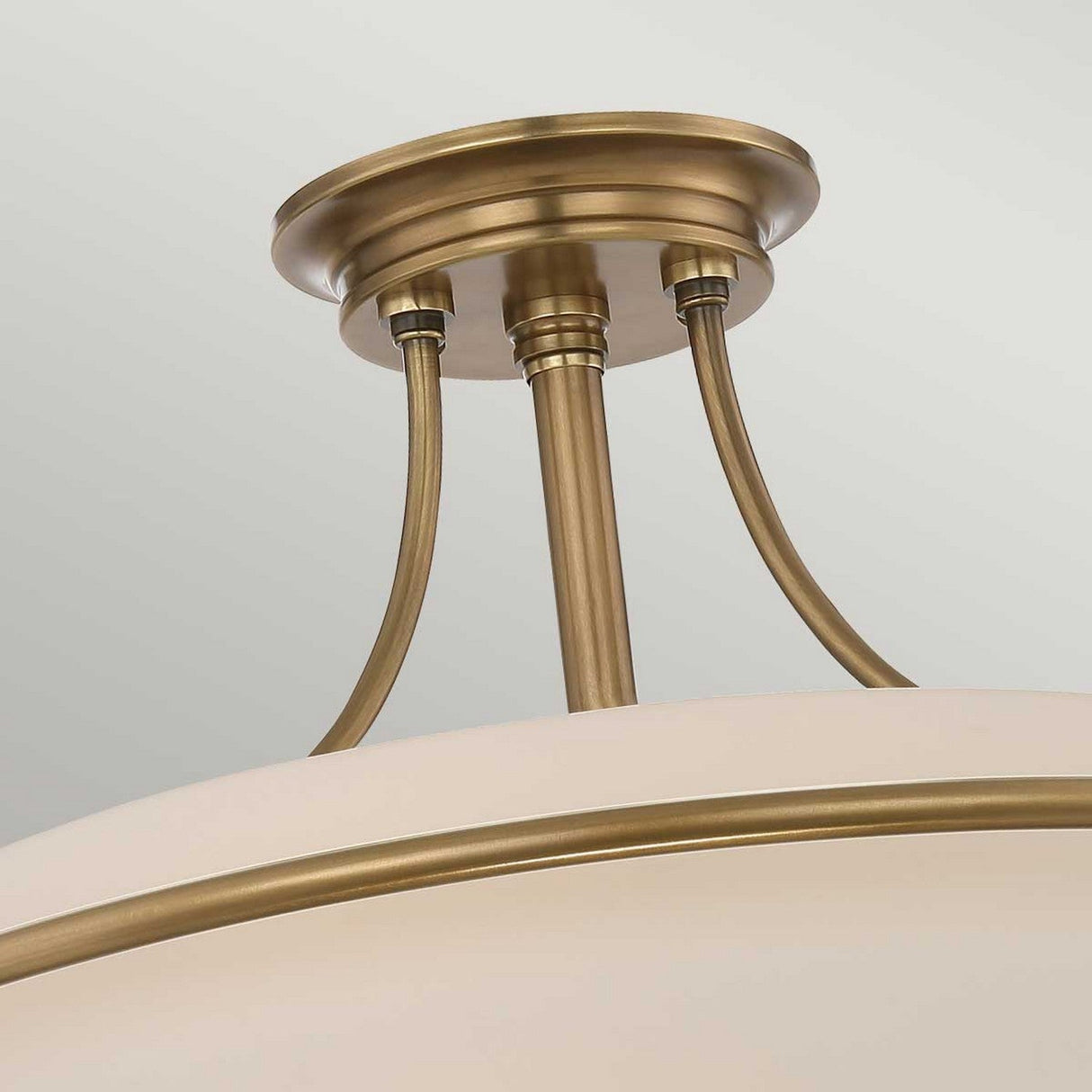 A close-up of the Griffin 4 Light Semi-Flush in weathered brass showcases its curved rods connecting to an opaque white shade. This fixture offers ambient lighting with a circular base, exuding both modern and vintage charm.