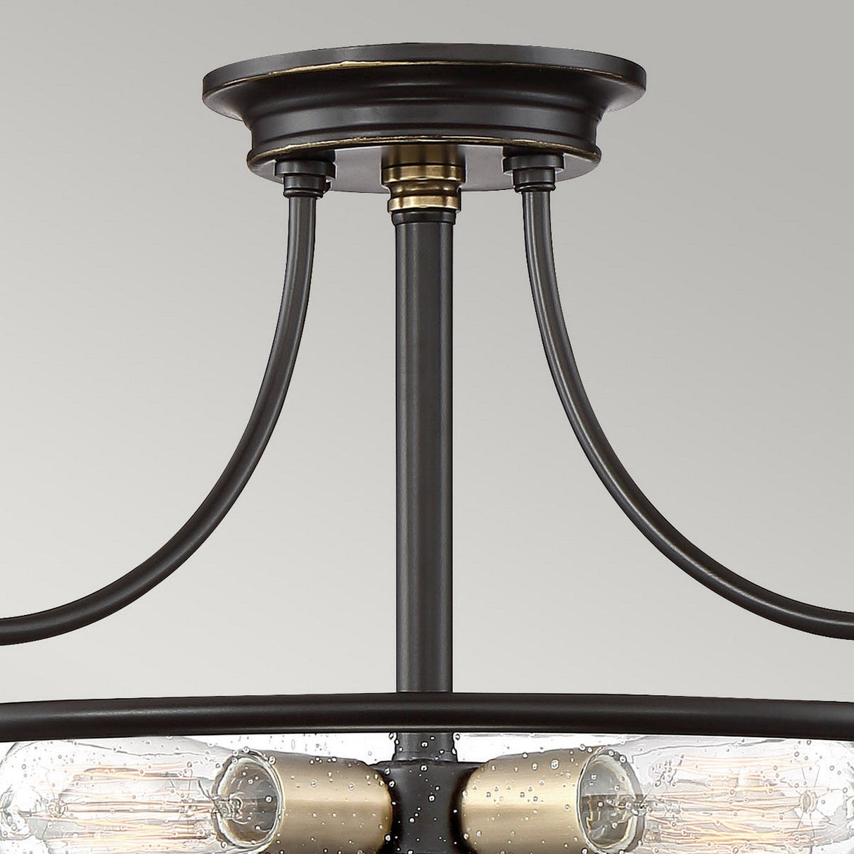 Close-up of the Griffin 3 Light Small Semi-Flush fixture, featuring a circular base with two curved supports. It showcases Palladian Bronze tones with clear glass shades that reveal bulb filaments, creating a modern and industrial look. The background is a plain gray.