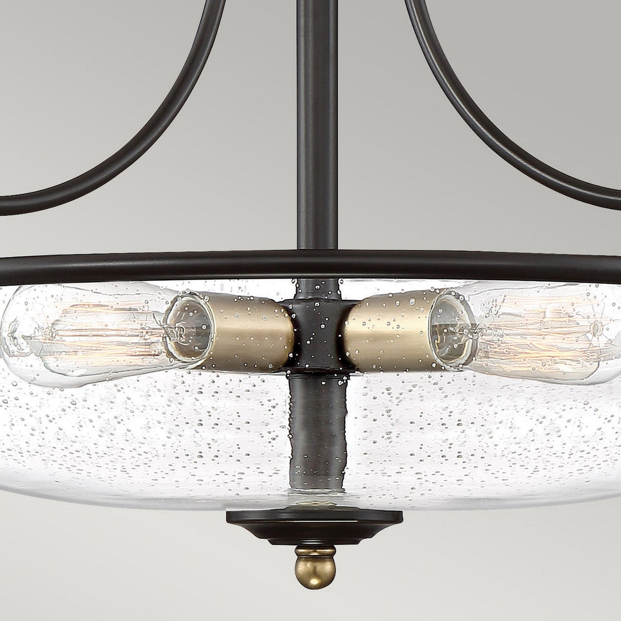 Close-up of the Griffin 3 Light Small Semi-Flush, showcasing exposed Edison bulbs nestled within a rounded clear glass shade dotted with tiny bubbles. The metallic frame is finished in Palladian Bronze and Weathered Brass accents, all set against a smooth, light gray backdrop.