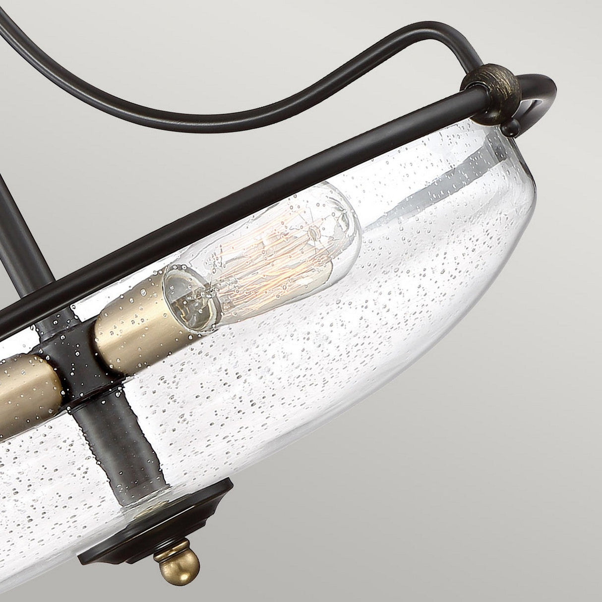 Close-up of the Griffin 3 Light Small Semi-Flush fixture highlighting an Edison-style bulb encased in clear, speckled glass. It boasts Palladian Bronze with Weathered Brass accents and curved lines against a neutral background, elevating the modern aesthetic with timeless elegance.