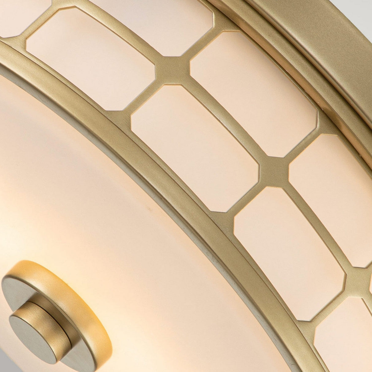 A close-up of the Guardian 2 Light Flush in natural brass reveals a geometric lamp fixture with a hexagonal pattern on frosted glass. The bathroom lighting design elegantly combines curved lines and straight edges within its metal cage for a modern look.