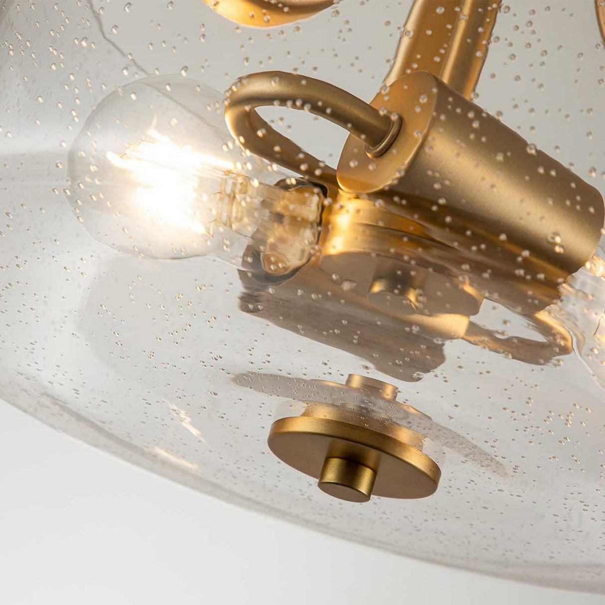 A close-up of the Hollister 2 Light Semi-Flush - Brushed Brass reveals its elegant design, featuring a brushed brass finish and a clear glass shade adorned with textured bubbles. The bulb inside casts a warm glow, enhancing its contemporary aesthetic.