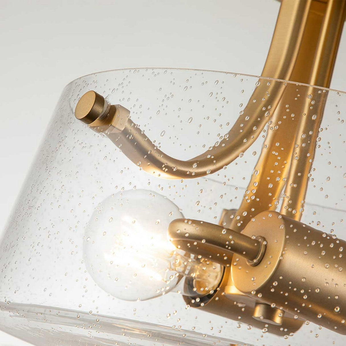 Close-up of the Hollister 2 Light Semi-Flush - Brushed Brass fixture, showcasing its clear glass shade adorned with delicate bubbles. The brushed brass finish complements the glowing bulb inside, giving it a contemporary and elegant look.