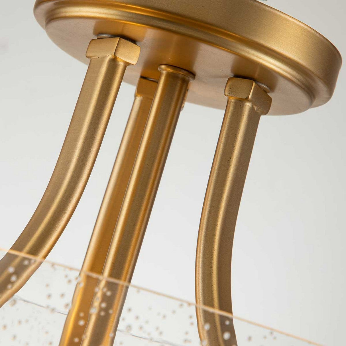 A close-up of the Hollister 2 Light Semi-Flush in brushed brass reveals three elegantly curved metal rods extending from the base, complemented by a glass component adorned with delicate bubbles for added sophistication.