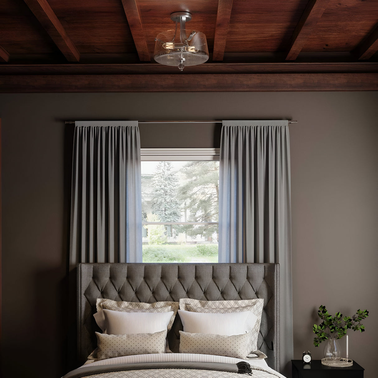 A cozy bedroom showcasing a tufted gray headboard, several pillows, and a neatly made bed. Light gray curtains gracefully frame a window overlooking lush greenery. A plant rests on the dark nightstand, illuminated by the warm glow of the Hollister 2 Light Semi-Flush - Polished Nickel with seeded glass shades.