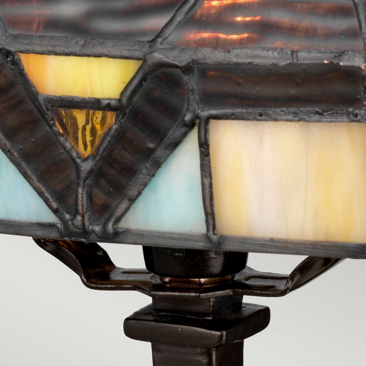 Close-up of the Holmes 1 Light Mini Tiffany Table Lamp in Vintage Bronze, featuring a stained glass shade with geometric patterns in shades of yellow, orange, brown, and blue. The dark vintage bronze framework complements the vibrant glass pieces, while the lamp base is partially visible.
