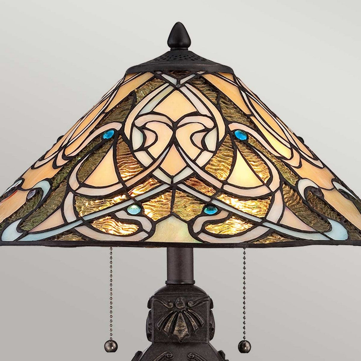 The India 2 Light Tiffany Table Lamp - Imperial Bronze features a captivating stained glass shade with an intricate, multicolored design. The lampshade showcases cream, amber, and blue patterns that beautifully complement the two pull chains and the elegant Imperial Bronze finish.
