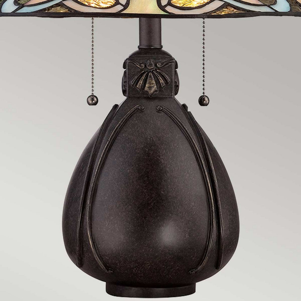 Close-up of the India 2 Light Tiffany Table Lamp in imperial bronze, showcasing its dark, rounded ornate base with a textured finish and decorative insect motifs near the top. Pull chains hang below the edge of its stained glass shade, set against a light gray background.