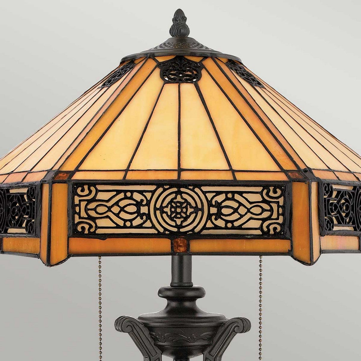 The Indus 2 Light Tiffany Table Lamp in Vintage Bronze features an amber glass shade adorned with a geometric pattern and ornate black metalwork, resting on a decorative vintage bronze base. The lampshade displays warm amber hues with intricate circular and linear designs, bringing elegance to any space.
