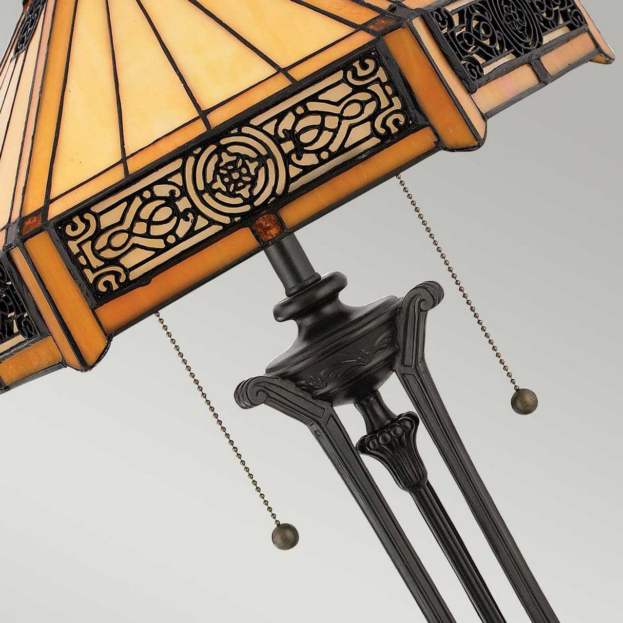 A close-up of the Indus 2 Light Tiffany Table Lamp - Vintage Bronze highlights its amber glass shade with a vintage-style design. The lamp's base features intricate metalwork in a vintage bronze finish, complemented by black accents, and two elegantly hanging pull chains.