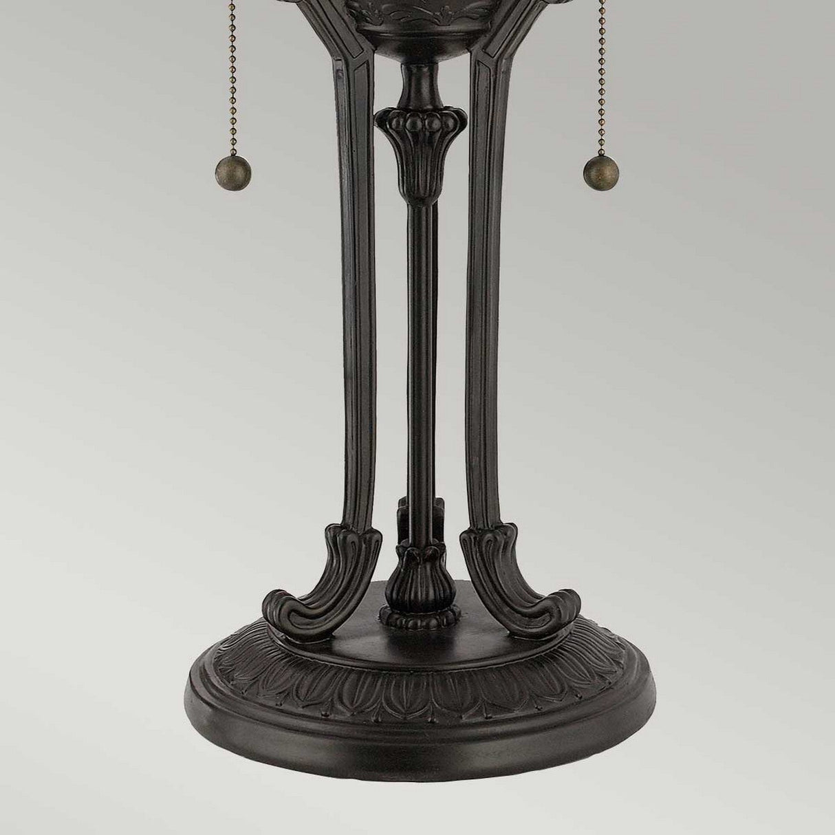 Capture the intricate detailing of the Indus 2 Light Tiffany Table Lamp's ornate base, adorned with elegant carvings in vintage bronze. It features two pull chains with small round weights and is beautifully topped with an amber glass shade. The subtle contrast against a plain light gray background enhances its timeless appearance.