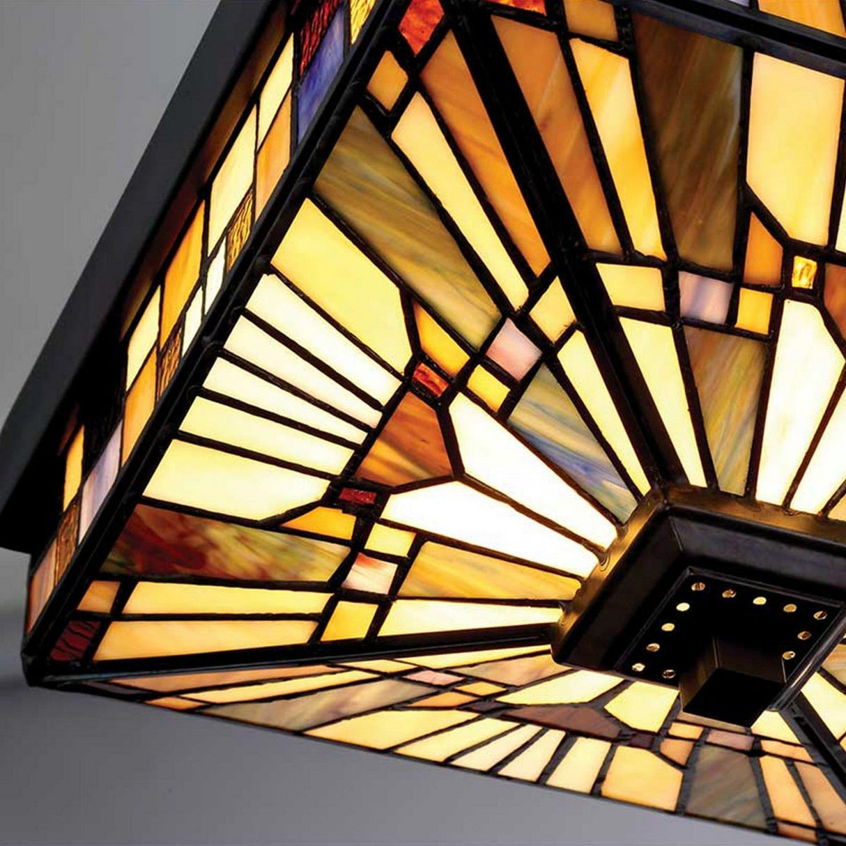 Detailed view of the Inglenook Outdoor 2 Light Tiffany Flush - Valiant Bronze, featuring a geometric stained glass design in yellow, amber, and red hues. It creates a sunburst pattern that casts a warm glow, with a dark frame that accentuates the vibrant colors.