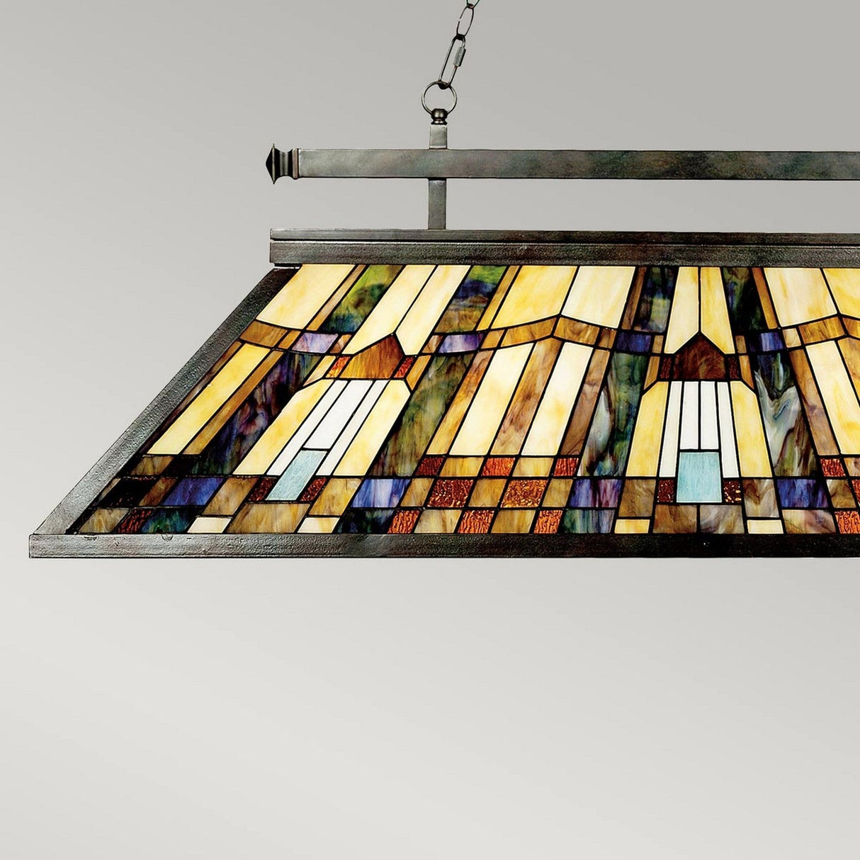 The Inglenook 3 Light Tiffany Island Chandelier in Valiant Bronze features a stunning stained glass shade with an intricate geometric pattern in shades of yellow, orange, brown, and blue. Its valiant bronze finish elegantly frames the light as it hangs from a chain against a neutral backdrop.