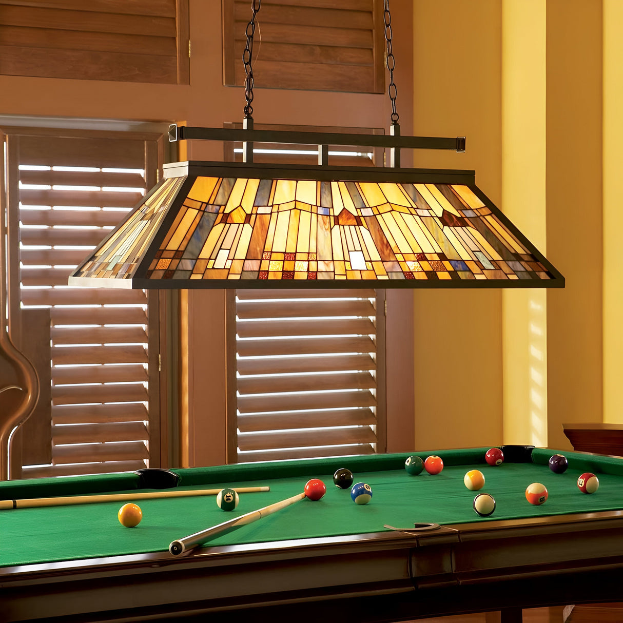 A billiards table with colored balls and cues sits beneath the Inglenook 3 Light Tiffany Island Chandelier in Valiant Bronze, complete with a stained glass shade. The room, accentuated by wooden shutters and warm yellow walls, exudes an inviting ambiance.