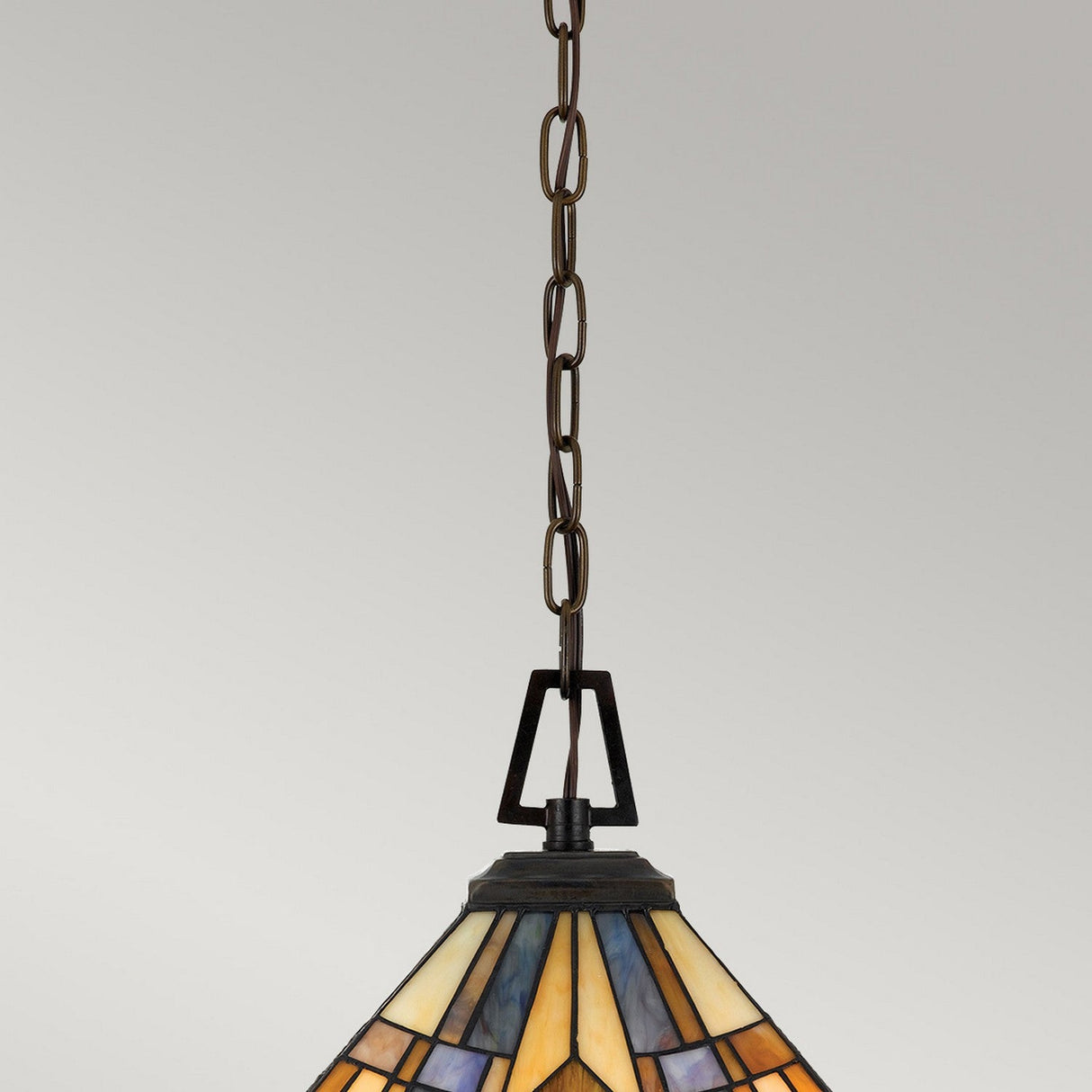 The Inglenook 3 Light Small Tiffany Pendant in Valiant Bronze elegantly hangs from the ceiling, with its chain supporting a stained glass shade that displays geometric patterns in shades of blue, amber, and cream.