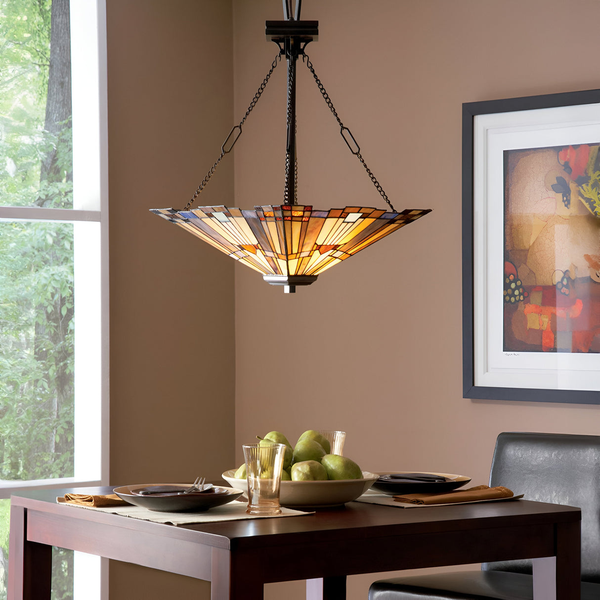 A dining area with a square wooden table set for two, featuring plates, glasses, and pears. Above the table hangs an Inglenook 3 Light Large Tiffany Pendant in Valiant Bronze with a stained glass shade. A window with a view of greenery and framed abstract art adorns the taupe-colored walls.