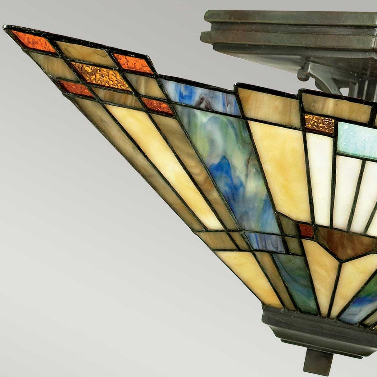 A close-up of the Inglenook 2 Light Tiffany Semi-Flush in Valiant Bronze showcases a stained glass shade featuring geometric patterns. Various shades of amber, blue, and orange glass pieces are meticulously arranged in an angular design, exuding a vintage and artistic charm.