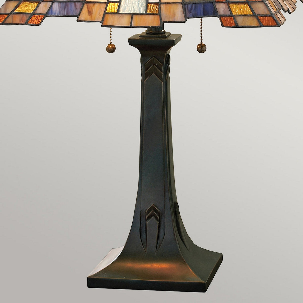 The Inglenook 2 Light Table Lamp in Valiant Bronze features a square base with a slender, elegantly carved column. Its lampshade displays a geometric art glass design in warm amber, purple, and cream tones. Two charming pull chains hang gracefully from the shade, enhancing this exquisite piece.
