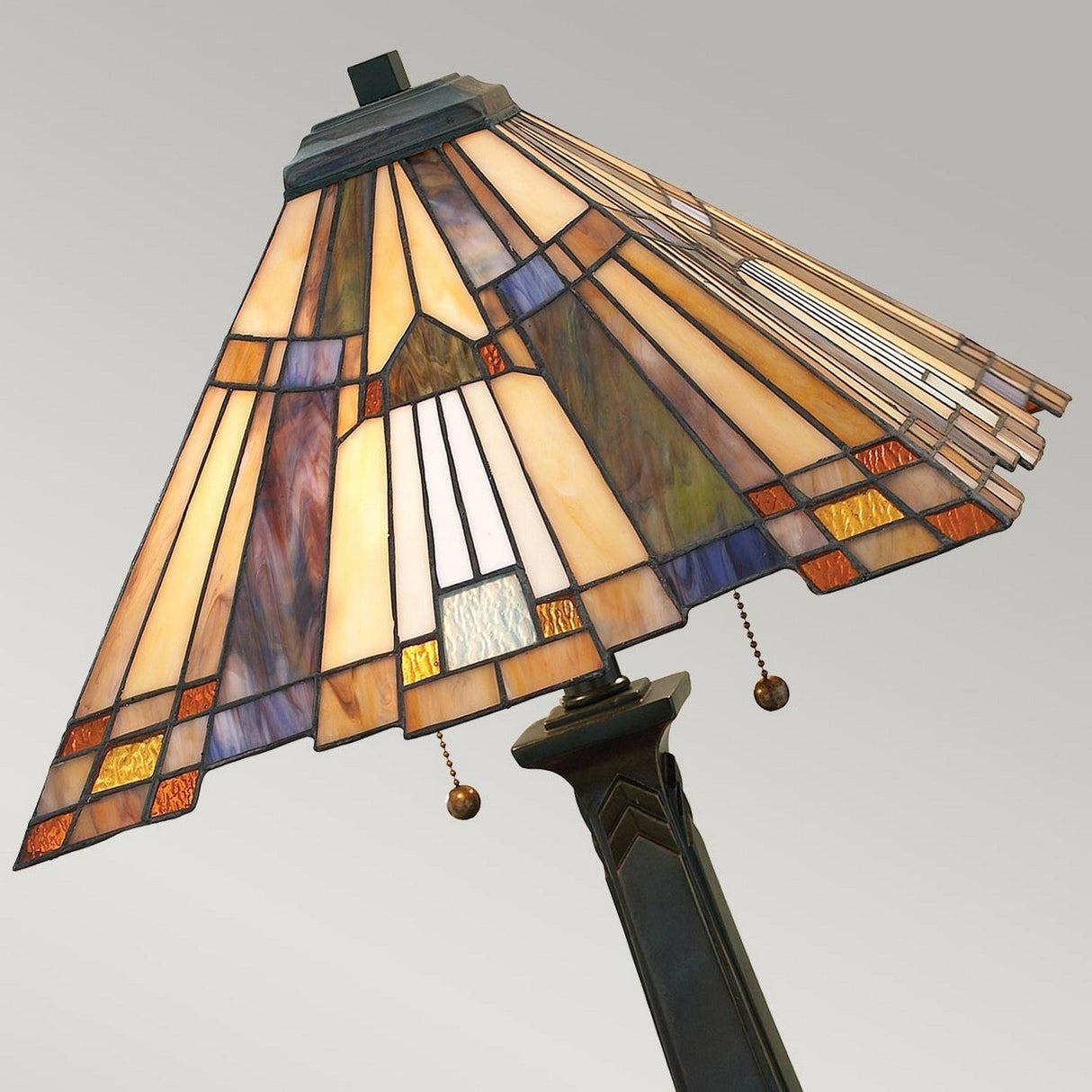 A close-up of the Inglenook 2 Light Table Lamp's stained glass lampshade, showcasing geometric patterns in orange, beige, and white hues. The lamp features a valiant bronze angular metal base with pull chains against a plain light gray background.