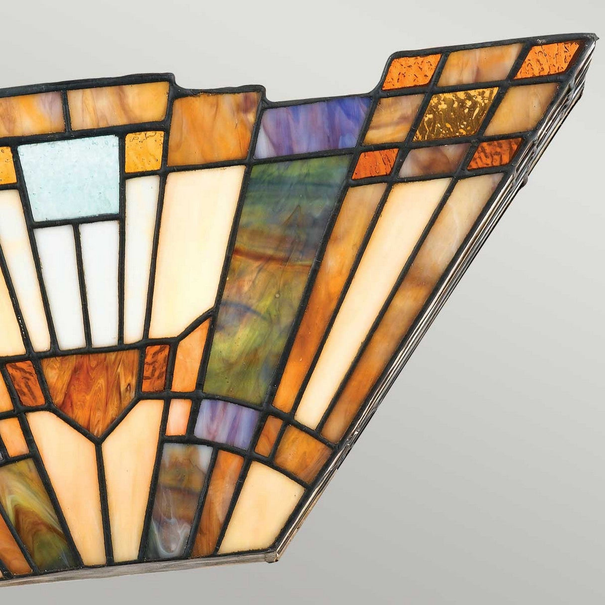 A close-up of the Inglenook 2 Light Tiffany Wall Light in Valiant Bronze showcases a stained glass panel with geometric patterns in shades of amber, brown, purple, and blue. Inspired by classic Arts & Crafts lighting, the design features elongated triangular shapes and a symmetrical arrangement, all framed by steel outlining each segment.