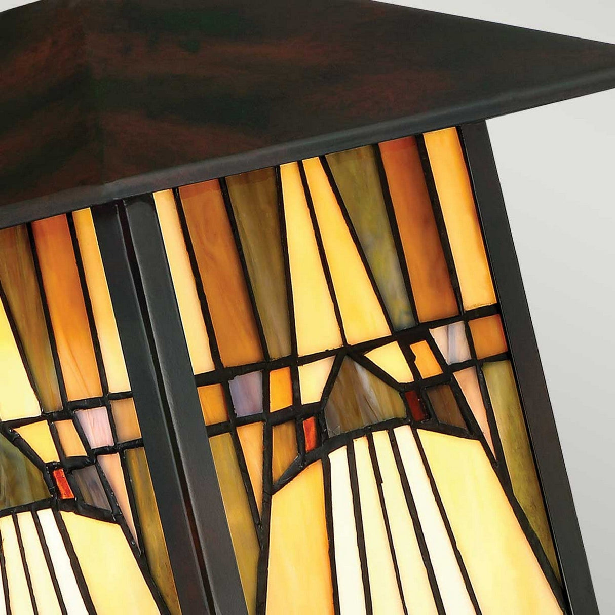 A detailed view of the Inglenook Outdoor 1 Light Medium Wall Lantern in Valiant Bronze showcases its Art Deco-inspired stained glass shade, adorned with geometric patterns in warm amber, yellow, and orange hues. The black outlines and subtle red accents reflect the Arts & Crafts style typical of outdoor wall lanterns.