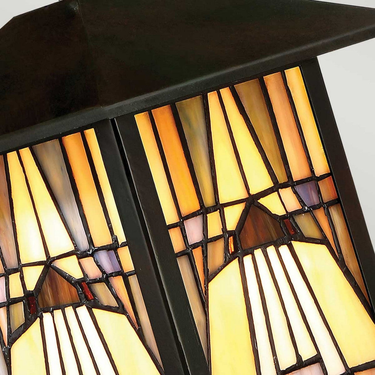 Close-up of the stained glass shade on the Inglenook Outdoor 1 Light Small Wall Lantern in Valiant Bronze. This design showcases a geometric pattern with beige, brown, and yellow tones framed by black metal, evoking a warm and inviting glow typical of the Arts & Crafts style.