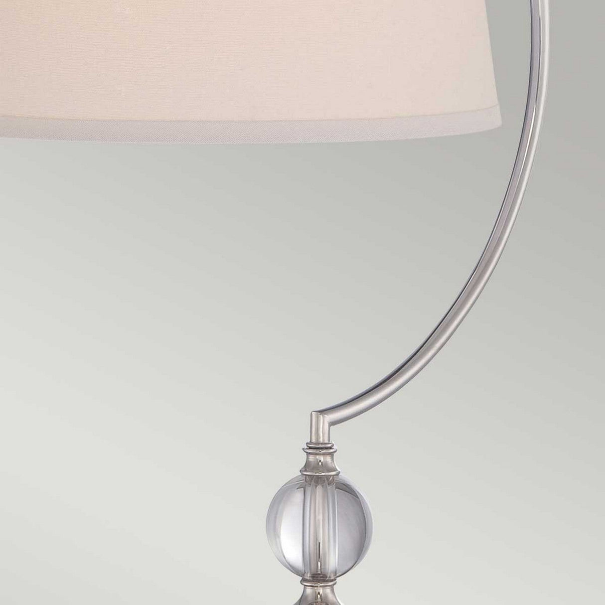 A close-up of the Jenkins 1 Light Table Lamp showcases its sleek polished nickel curved stand, adorned with a decorative glass sphere near the base, and topped with a light beige lampshade. Set against a neutral gray backdrop, this piece epitomizes contemporary decor.