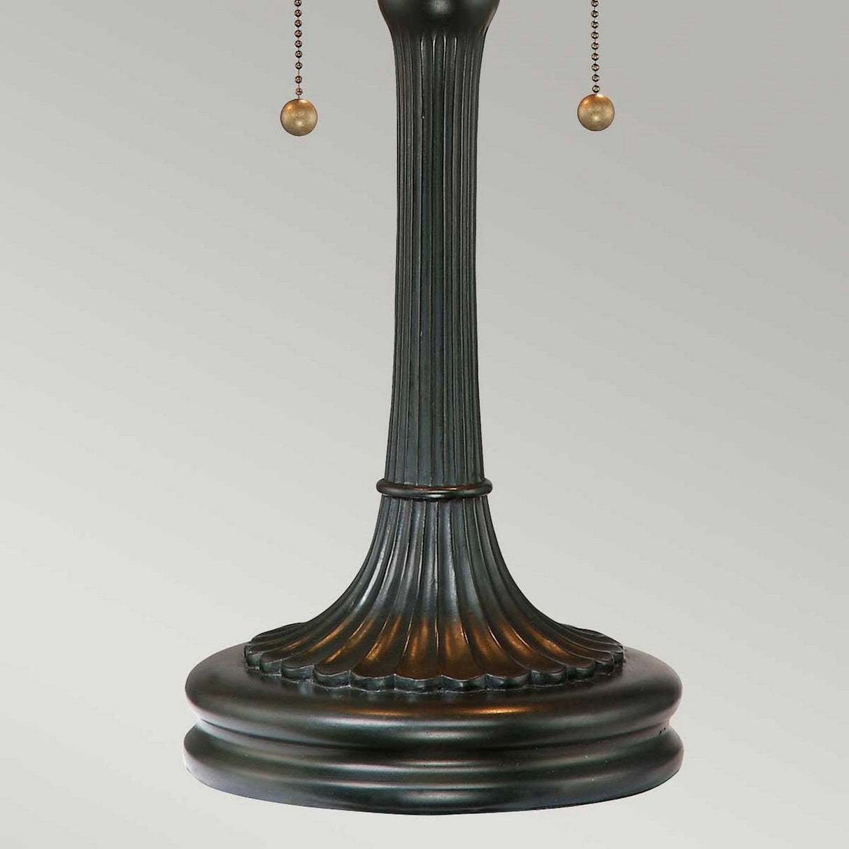 A close-up of the Kami 2 Light Tiffany Table Lamp - Vintage Bronze reveals an antique lamp base with a ribbed design and two hanging pull chains ending in small spheres. The vintage bronze base, with its weathered finish, echoes the charm of a classic Tiffany table lamp, all set against a light gray background.
